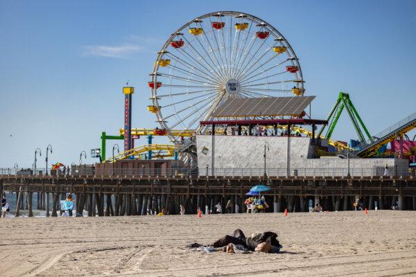 A homeless individual sleeps on the beach with the famed Ferris wheel at the Pacific Park in the background in Santa Monica, Calif., on Dec. 8, 2023. (John Fredricks/The Epoch Times)