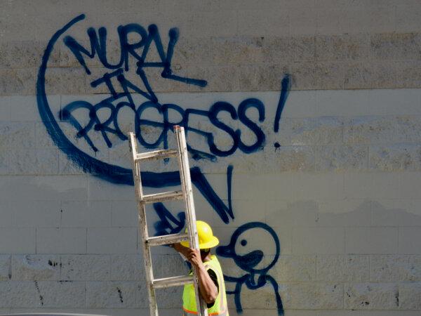 After the Beautification Council cleared a wall of graffiti, Emanuel Caprio paints a message signaling a mural is coming soon in Oakland, Calif., on May 2, 2024. (Travis Gillmore/The Epoch Times)