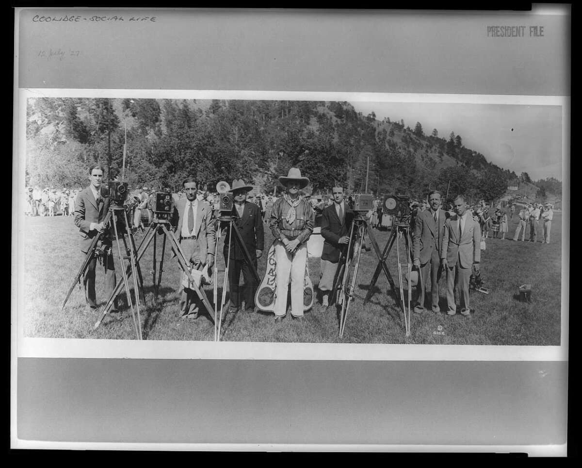 President Coolidge (C) in cowboy outfit, standing in field with photographers; mountain in background, 1927. Library of Congress. (Public Domain)