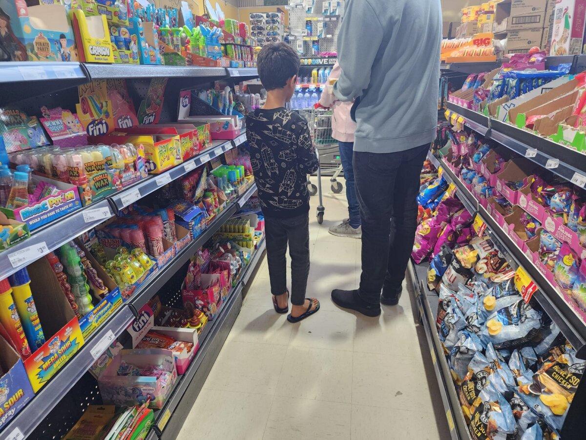 A child stands next to his father in a checkout aisle that is filled with colorfully packaged junk food marketed at children in a supermarket in Albany, Australia, on Sept. 7, 2024. (Susan Mortimer/The Epoch Times)
