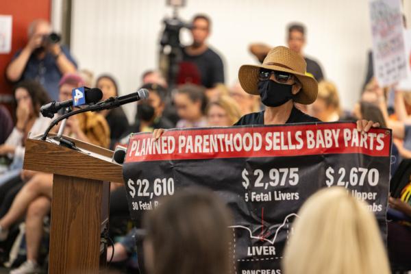 A woman holds up a sign from the podium of an Orange Unified School District meeting in Orange, Calif., on Aug. 17, 2023. (John Fredricks/The Epoch Times)