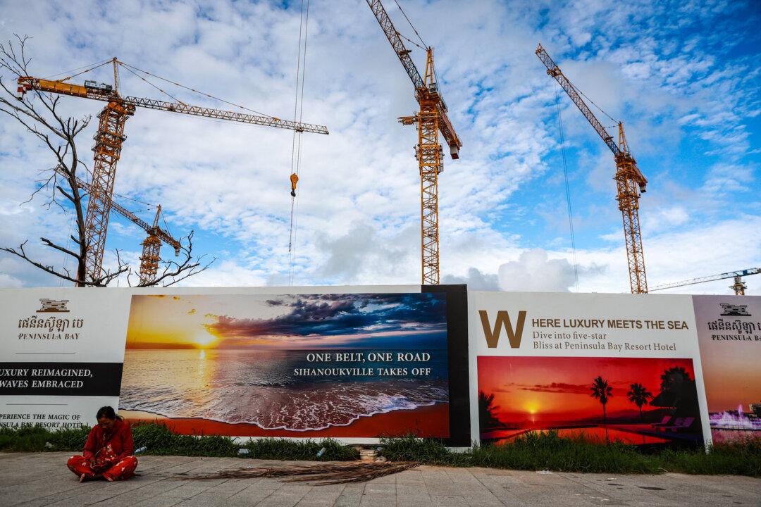 A woman sits near a billboard advertising China's Belt and Road Initiative in Sihanoukville, Cambodia, on July 1, 2024. Disguised as infrastructure development, the $1 trillion platform seizes other countries’ natural resources and expands its use of their ports for civil and military purposes. (Valeria Mongelli/Getty Images)