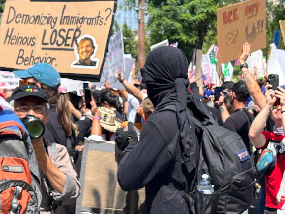 A masked protester wearing all black stands out from the colorful crowd at the No Kings protest at Los Angeles City Hall on June 14, 2025. (Brad Jones/The Epoch Times)