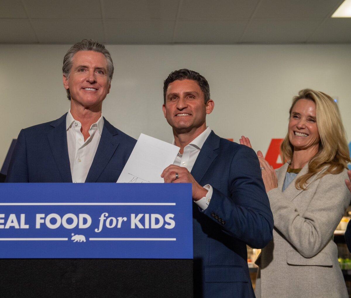 Gov. Gavin Newsom (L) signs AB 1264, a bill to phase out ultra-processed foods from schools, on Oct. 8, 2025. The bill's author, Assemblyman Jesse Gabriel (D-Encino) (C), holds the bill while Newsom's wife Jennifer Siebel Newsom applauds. (Gov. Gavin Newsom's Office)