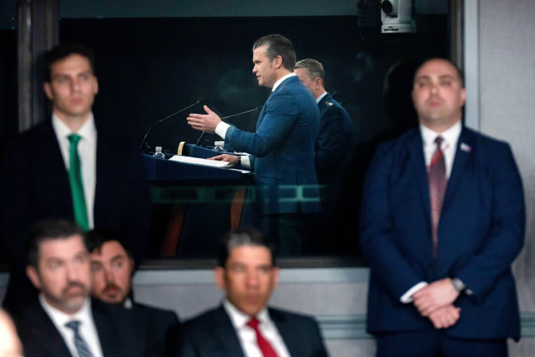 Staff members watch as Defense Secretary Pete Hegseth, reflected in a window, speaks during a press briefing at the Pentagon on March 2, 2026. (Mark Schiefelbein/AP Photo)