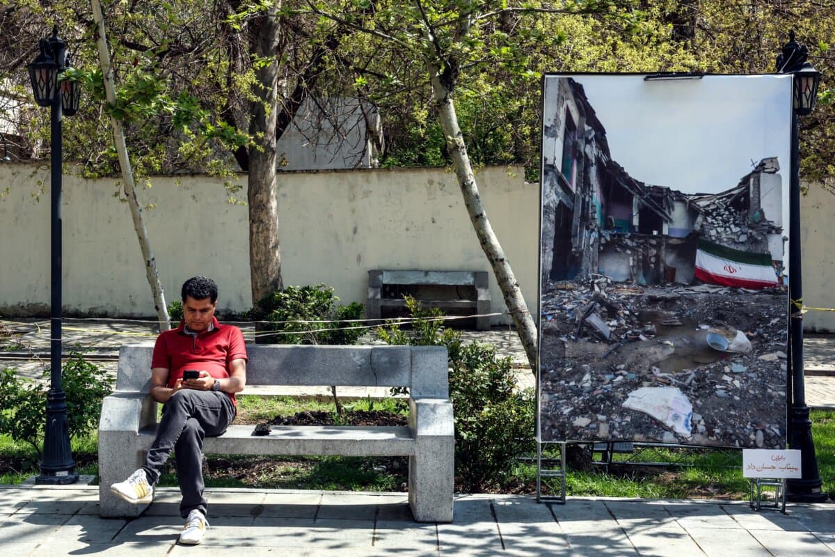 An Iranian man looks at his mobile phone as he sits near a large photograph depicting the current "Middle East war" in a Tehran park on April 16, 2026. (AFP via Getty Images)