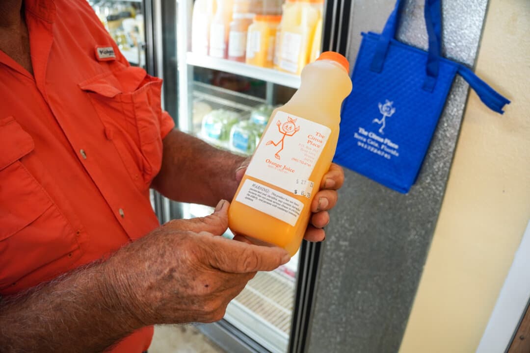(Top) A bin of Minneola tangelos can be seen at The Citrus Place in Terra Ceia, Florida, on May 21, 2026. Because of devastation caused by citrus greening, the fruit sold in the market is sourced from other farmers in the state. (Bottom) Lifelong citrus farmer Sidney Tillett shows his popular fresh-squeezed orange juice at The Citrus Place in Terra Ceia, Florida, on May 21, 2026. The orange was once the state's largest cash crop. (Jacob Burg/The Epoch Times)
