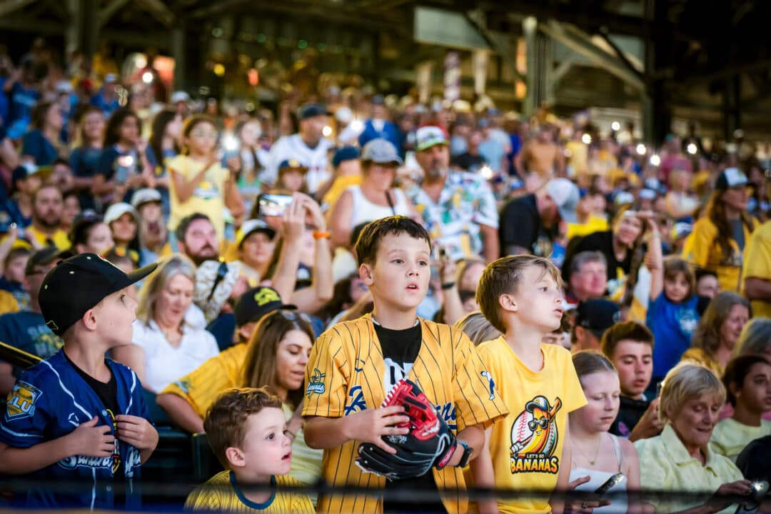 (Top) Pitcher Kyle Luigs of the Savannah Bananas winds up for the throw at Coors Field in Denver on Aug. 15, 2026. (Bottom Left) Young fans watch the game at Coors Field in Denver on Aug. 15, 2026. (Bottom Right) Savannah Bananas team members on the field in Denver on Aug. 15, 2026. (Madalina Kilroy/The Epoch Times)