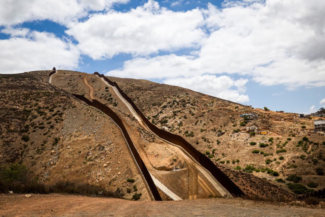 (Top) Construction vehicles navigate hilly terrain along the southern border wall outside San Diego on May 27, 2026. (Bottom) The southern border wall, seen from outside San Diego on May 27, 2026. (John Fredricks/The Epoch Times)
