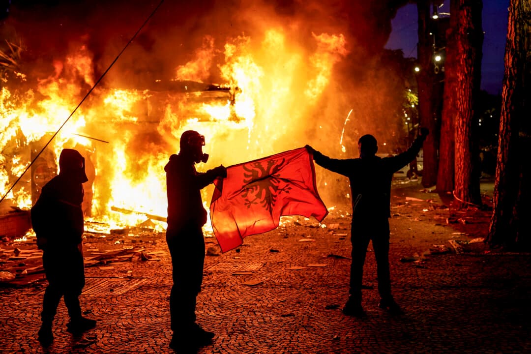 Protesters hold an Albanian flag in front of a burning kiosk during an anti-government demonstration in Tirana, Albania, on Oct. 15, 2026, as protests continued for a third consecutive day over university conditions and demands for the resignation of Albanian Prime Minister Edi Rama. (Adnan Beci/AFP via Getty Images)